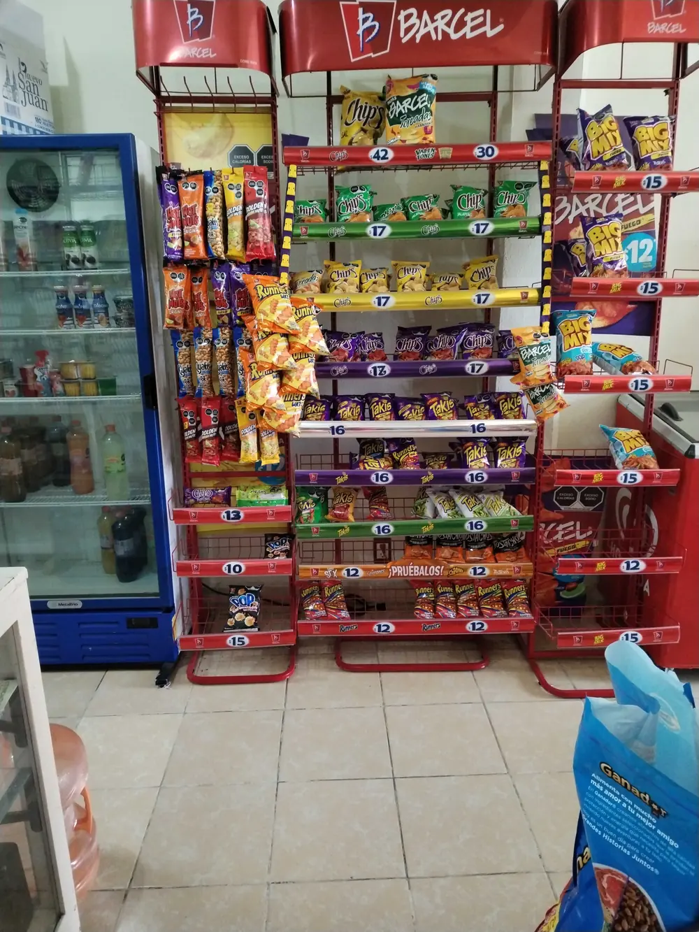 Branded snack rack beside a drinks cooler on the tiled floor of a small independent store.