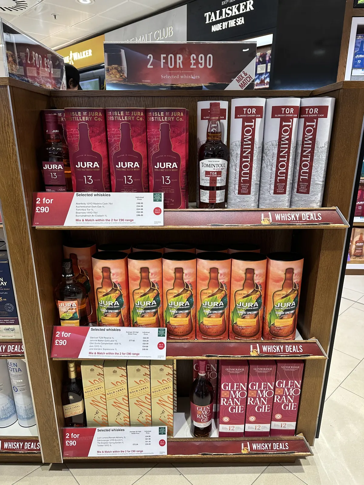 Whisky shelf in UK travel retail with Jura 13, Tomintoul and Glenmorangie bottles under 2 for £90 mix and match signage and shelf-edge price labels