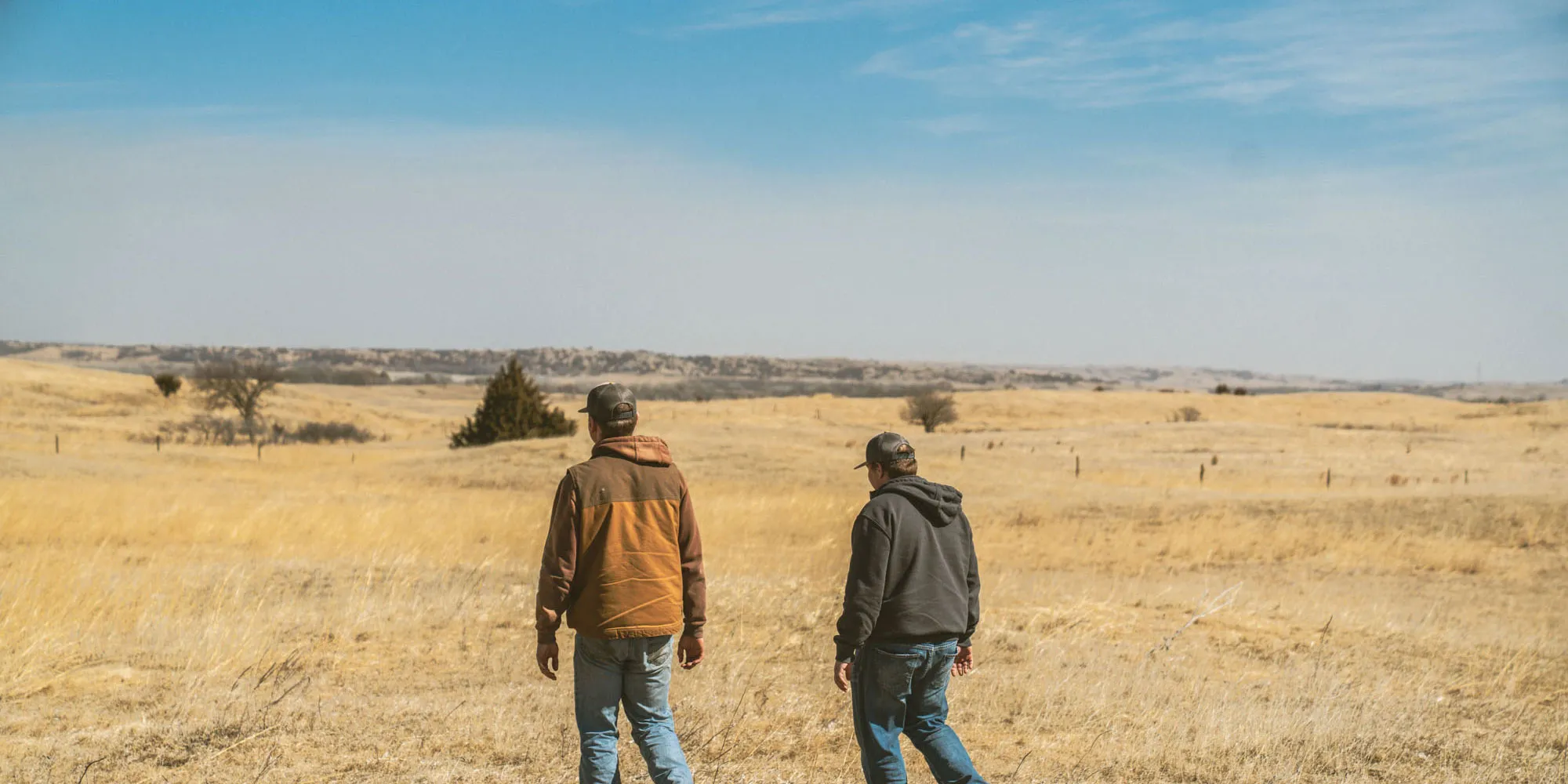 Two ranchers wearing caps and jackets walking across a vast dry grassy field under a blue sky.