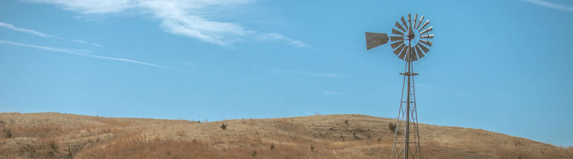 Old metal windmill standing on a dry, grassy hill under a clear blue sky.