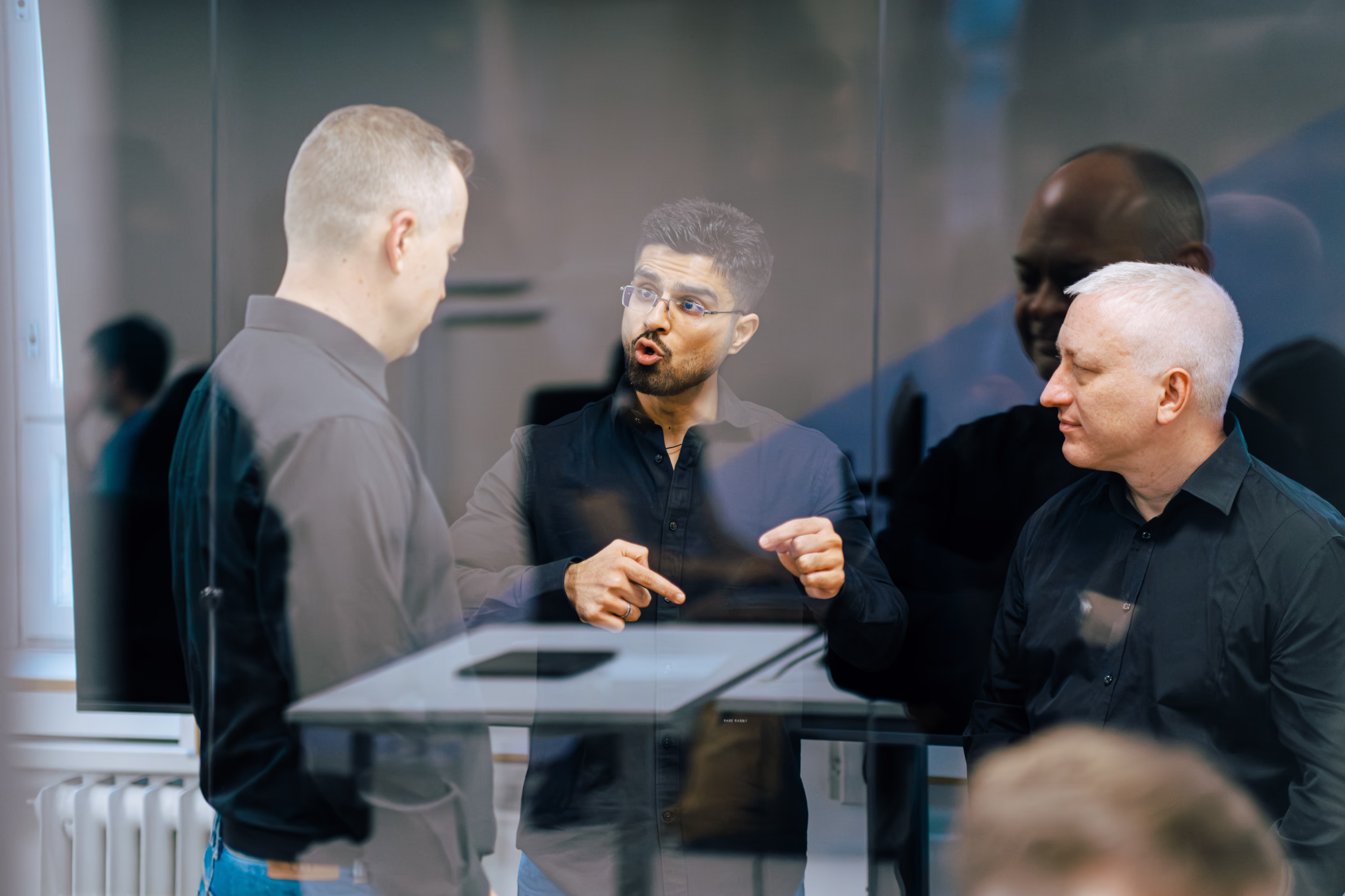Three men in black shirts talking inside an office with reflections on glass.
