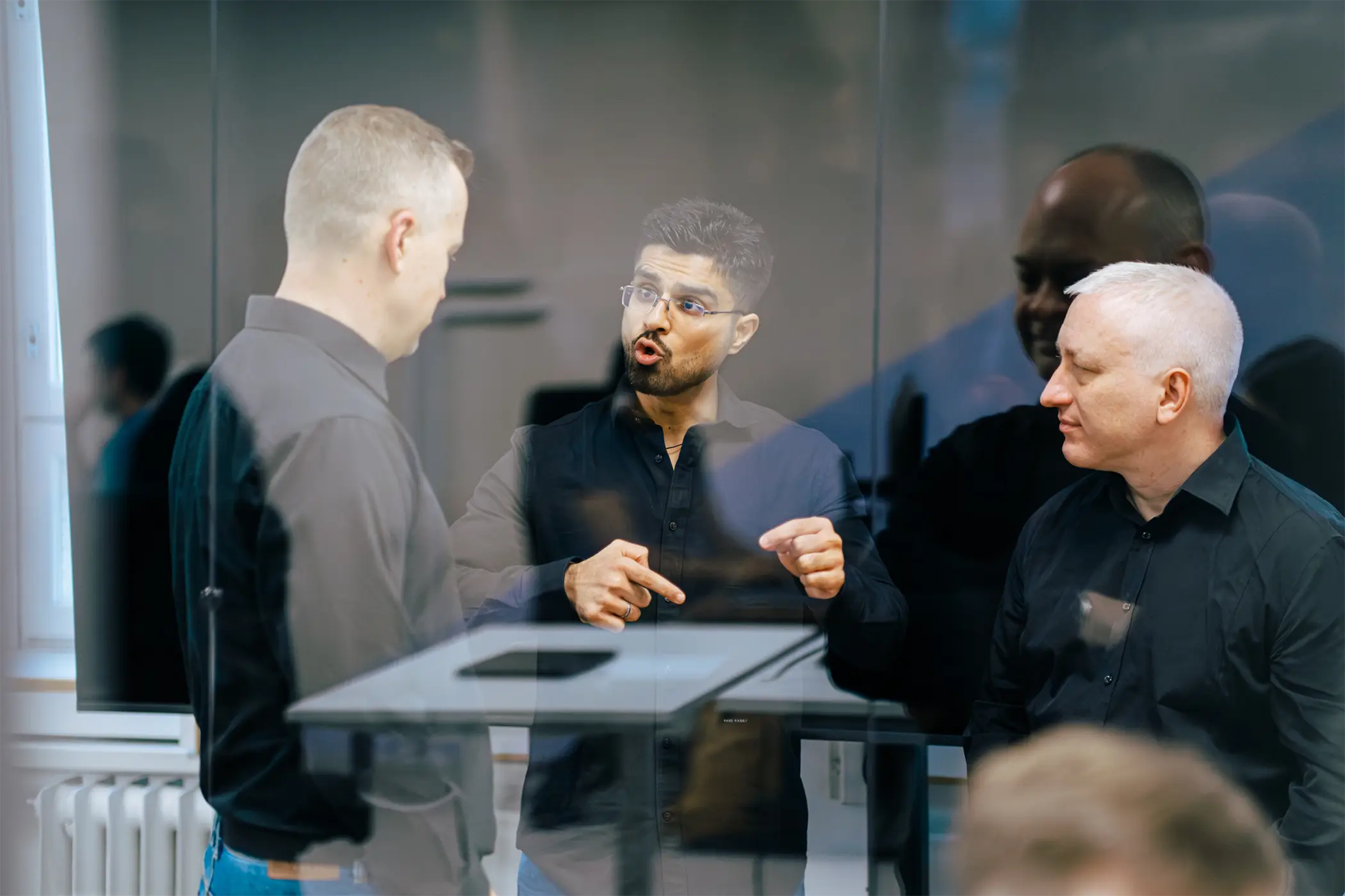 Three men in black shirts talking inside an office with reflections on glass.