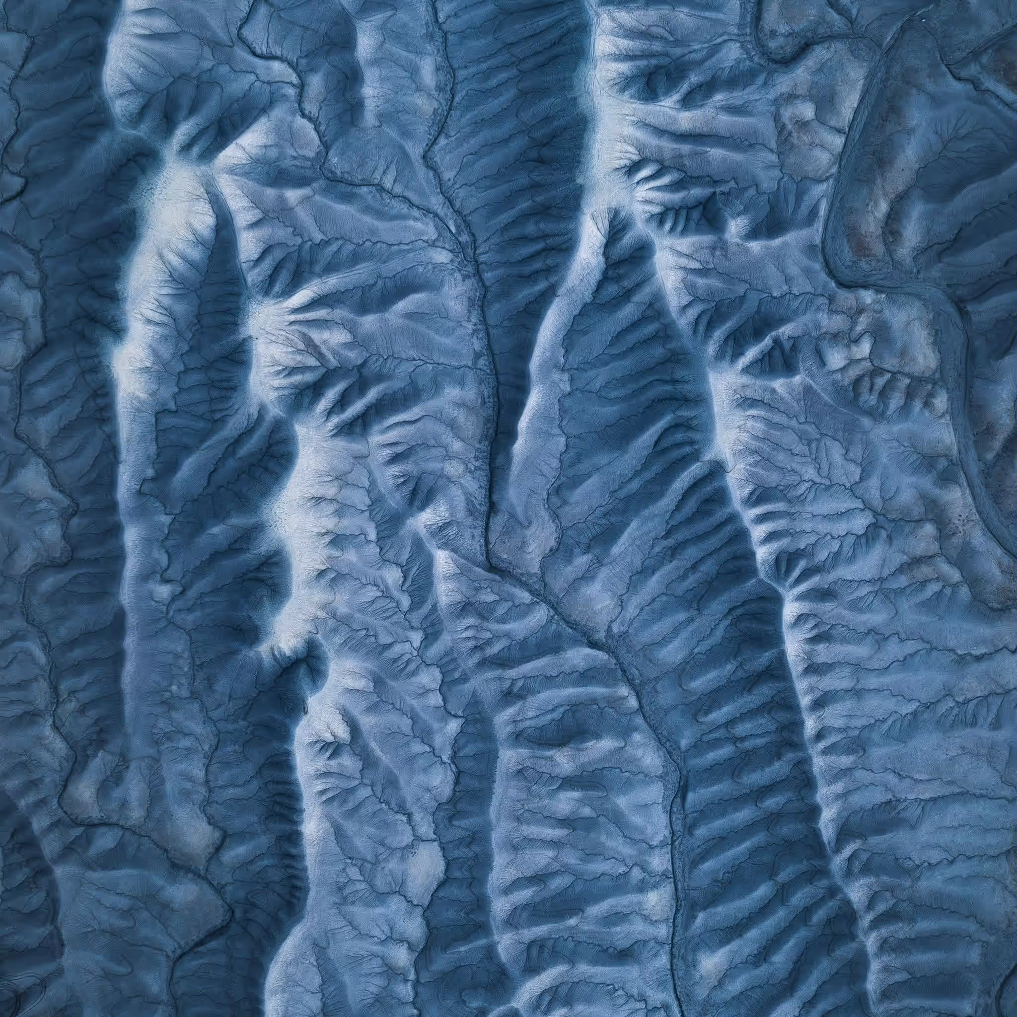 Aerial view of mountainous terrain with deep valleys and ridges in blue-gray tones.