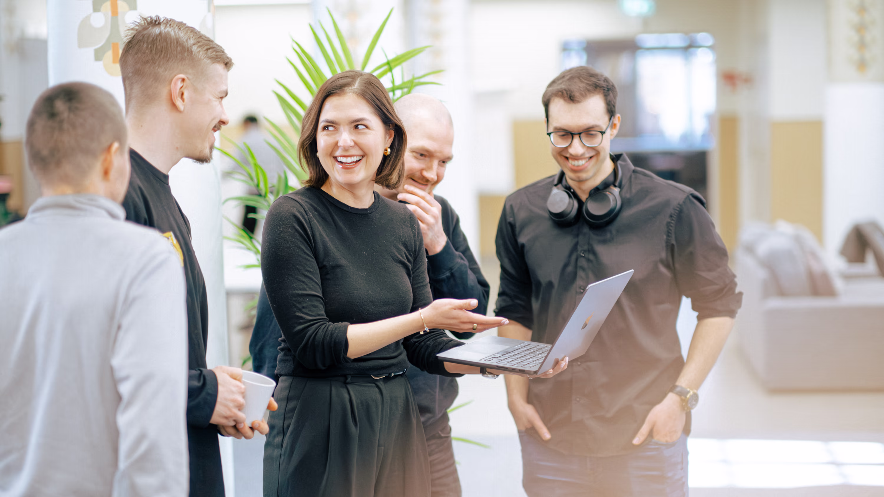 Group of five young professionals chatting and smiling around a laptop in a bright office space.
