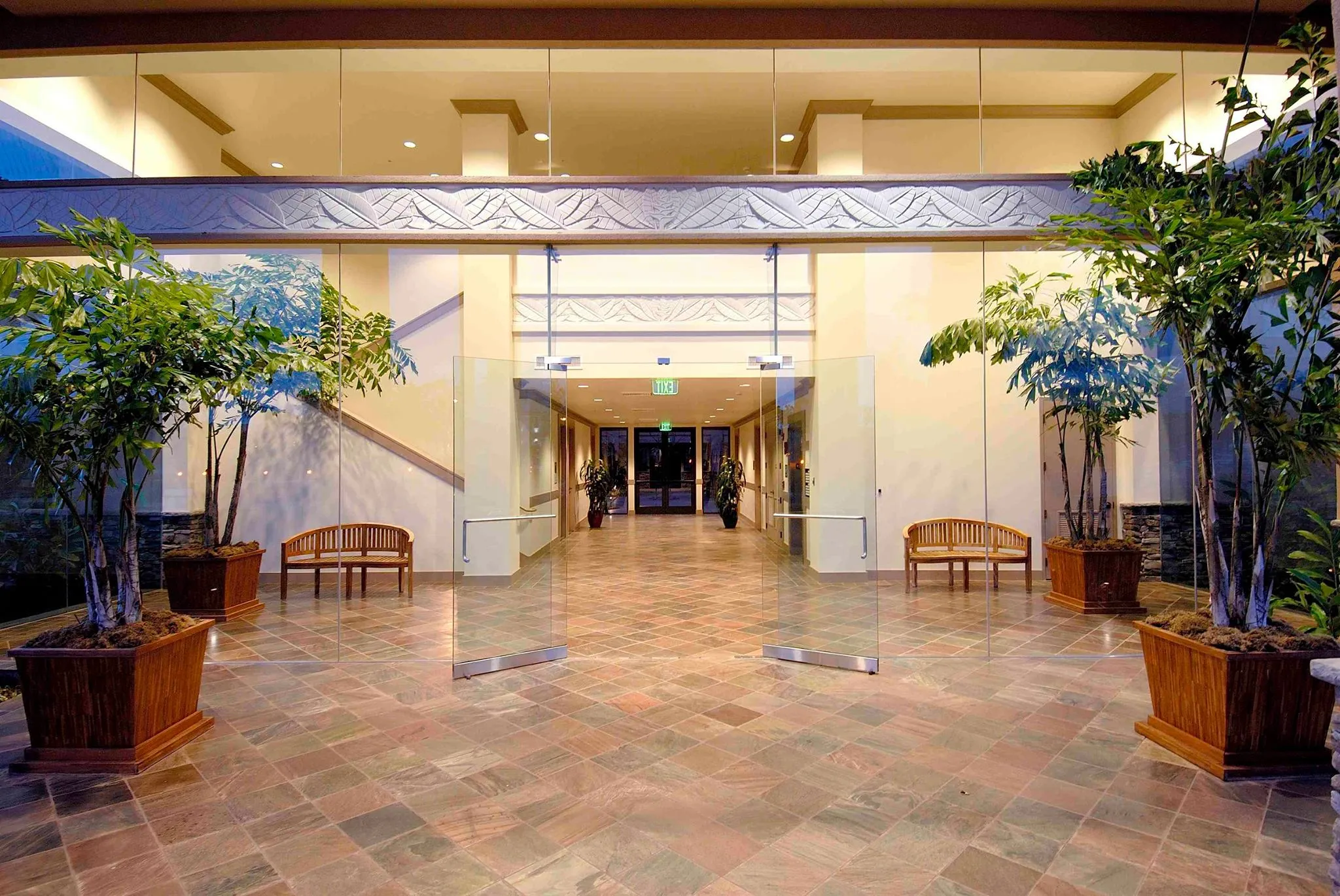 Modern building atrium with glass doors, wooden benches, potted plants, and decorative tilework.