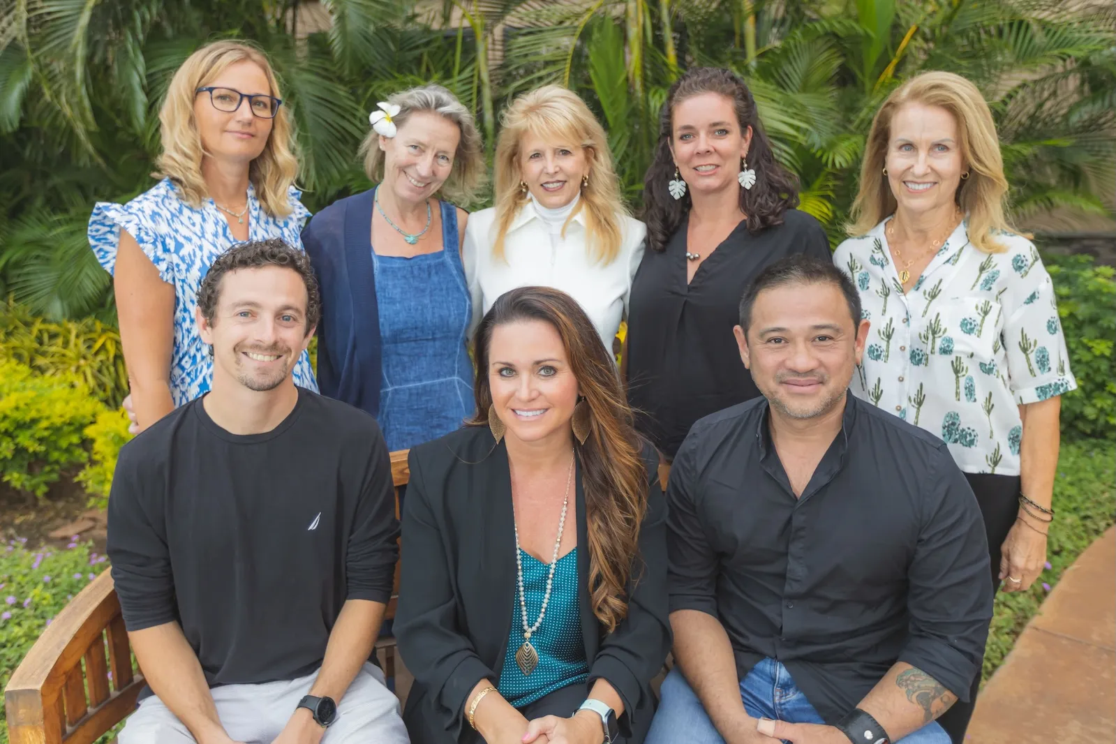 Group portrait of eight diverse people smiling outdoors with lush green tropical plants backdrop.