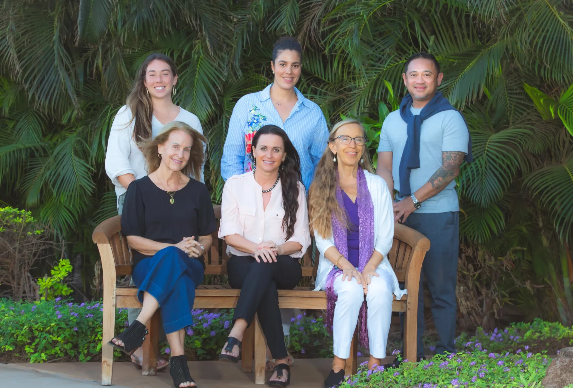 Six people posing together on wooden bench against tropical palm foliage backdrop