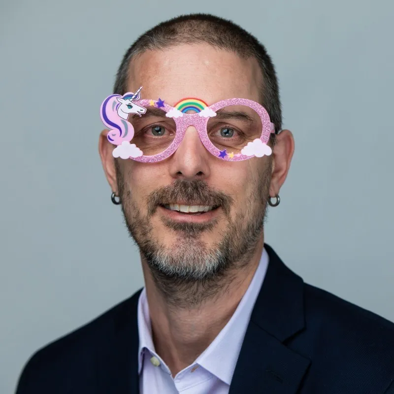 Middle-aged man with short hair and beard wearing a suit and glittery pink unicorn-themed novelty glasses.