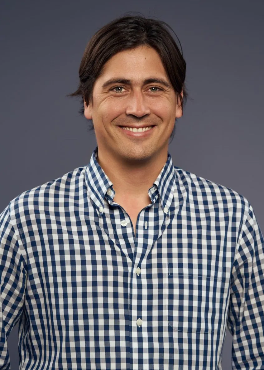 Smiling man with dark hair wearing a blue and white checkered button-up shirt against a plain gray background.