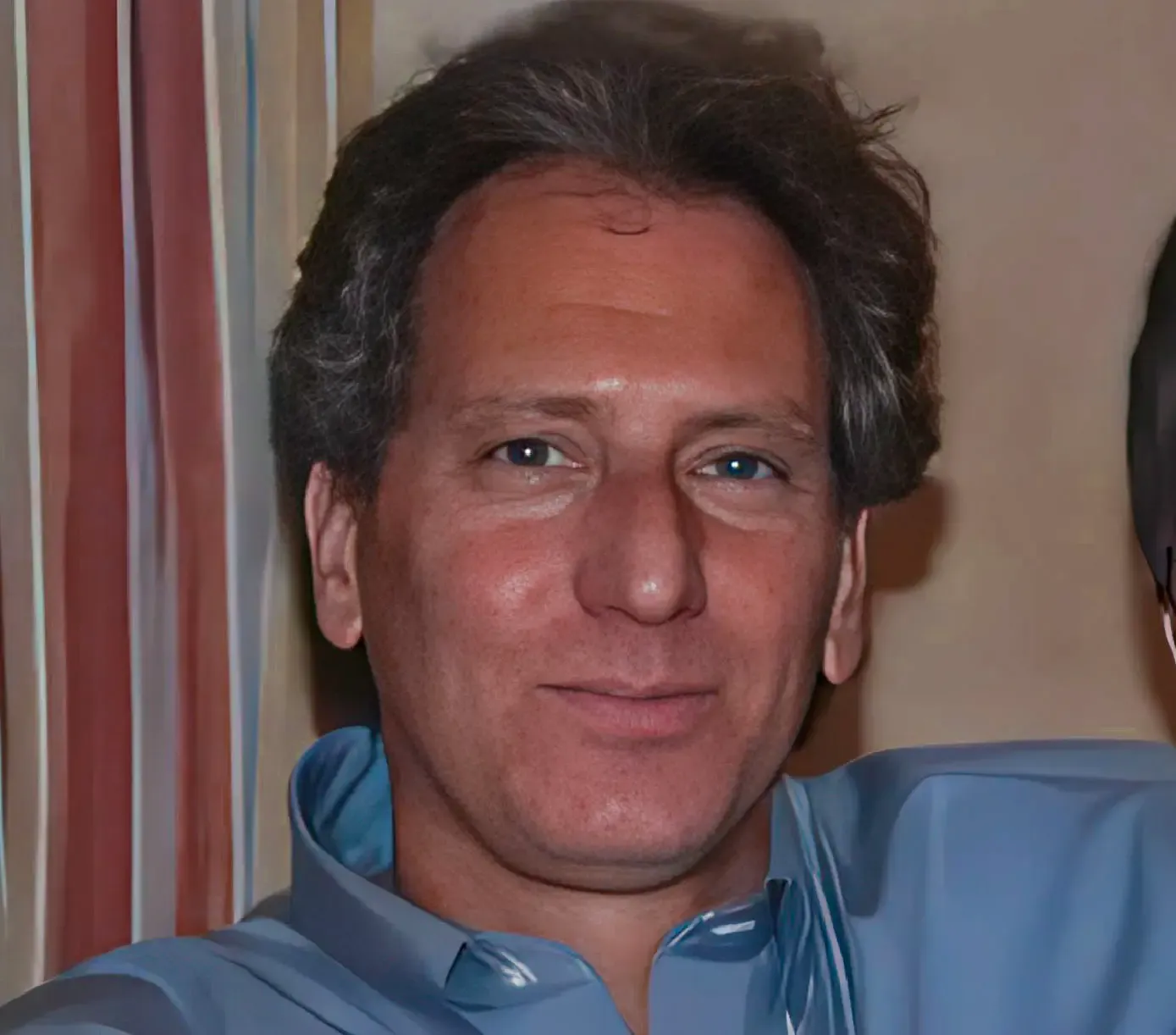 Middle-aged man with grayish hair wearing a light blue shirt smiling slightly indoors.