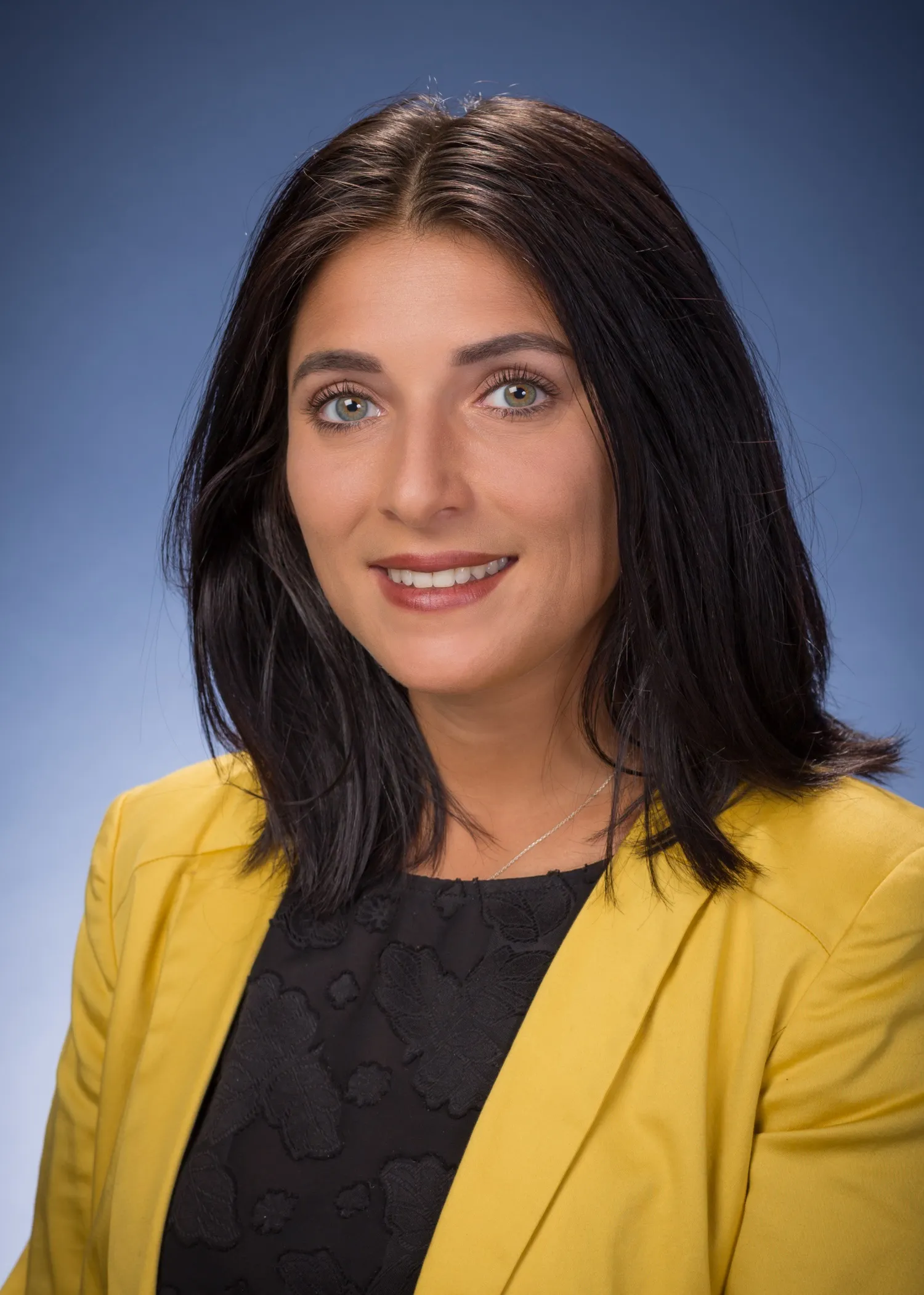Portrait of a woman with shoulder-length dark hair wearing a yellow blazer and black patterned top, smiling against a blue gradient background.