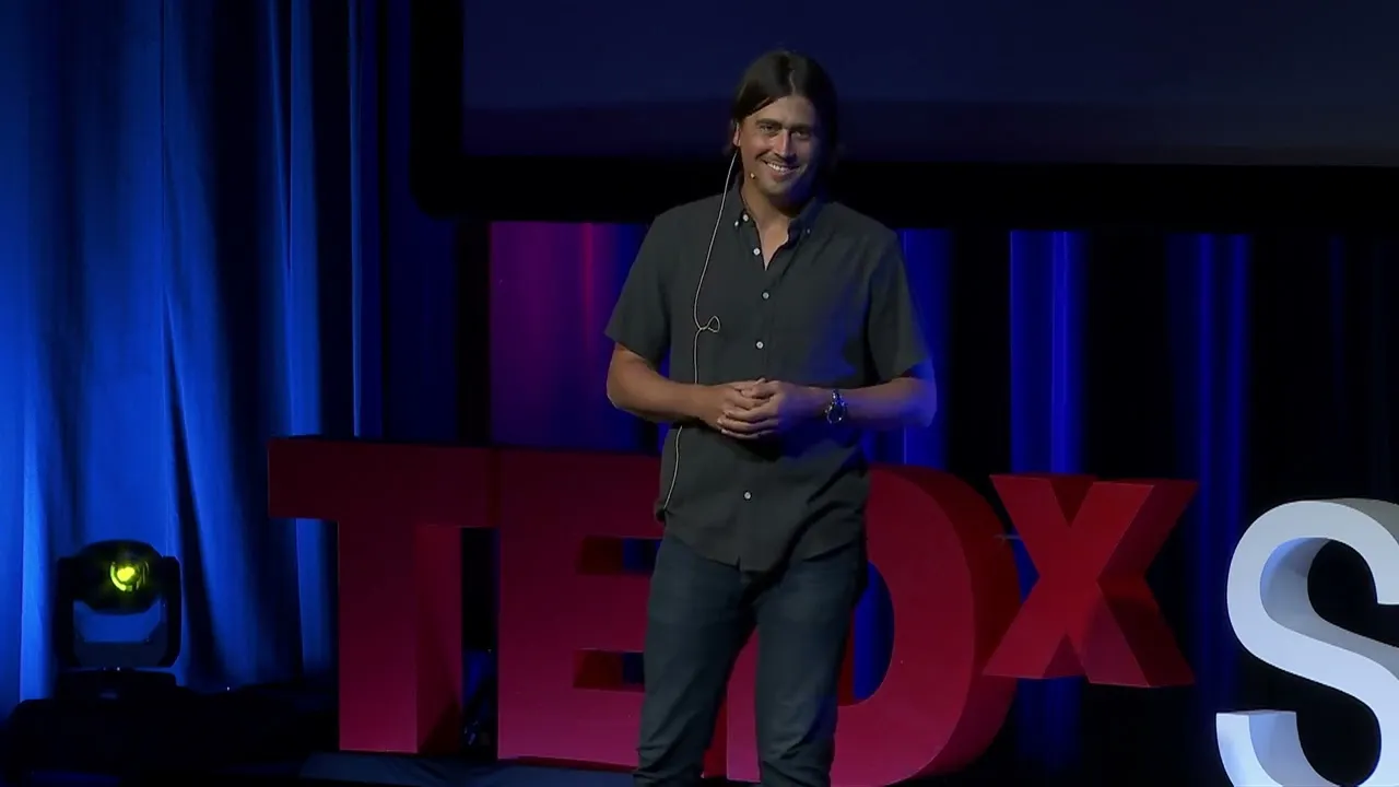 Man with long hair in a dark shirt speaking on stage at a TEDx event with large red TEDx letters behind him.