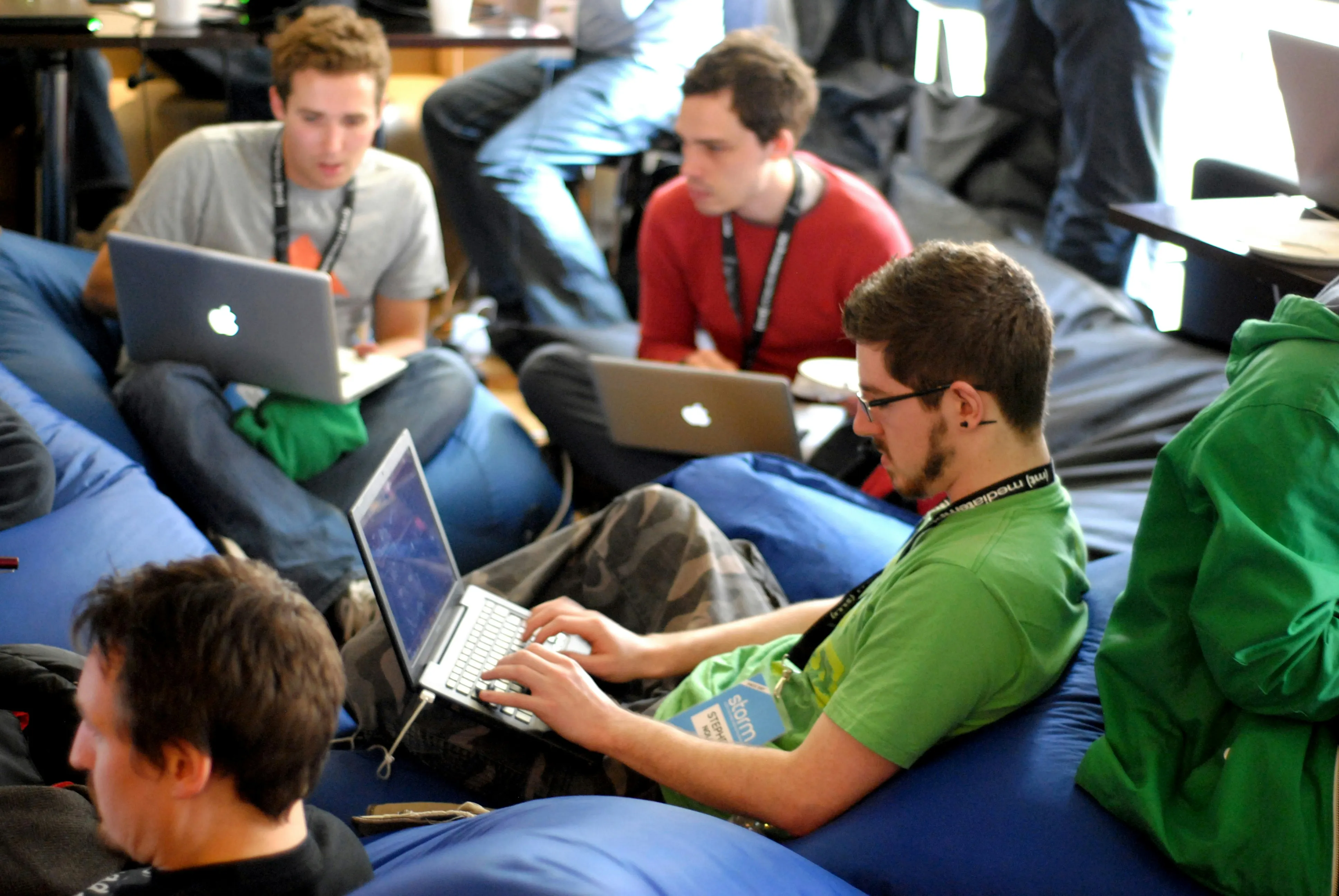 Group of young men sitting on blue bean bags working on laptops in a casual indoor setting.