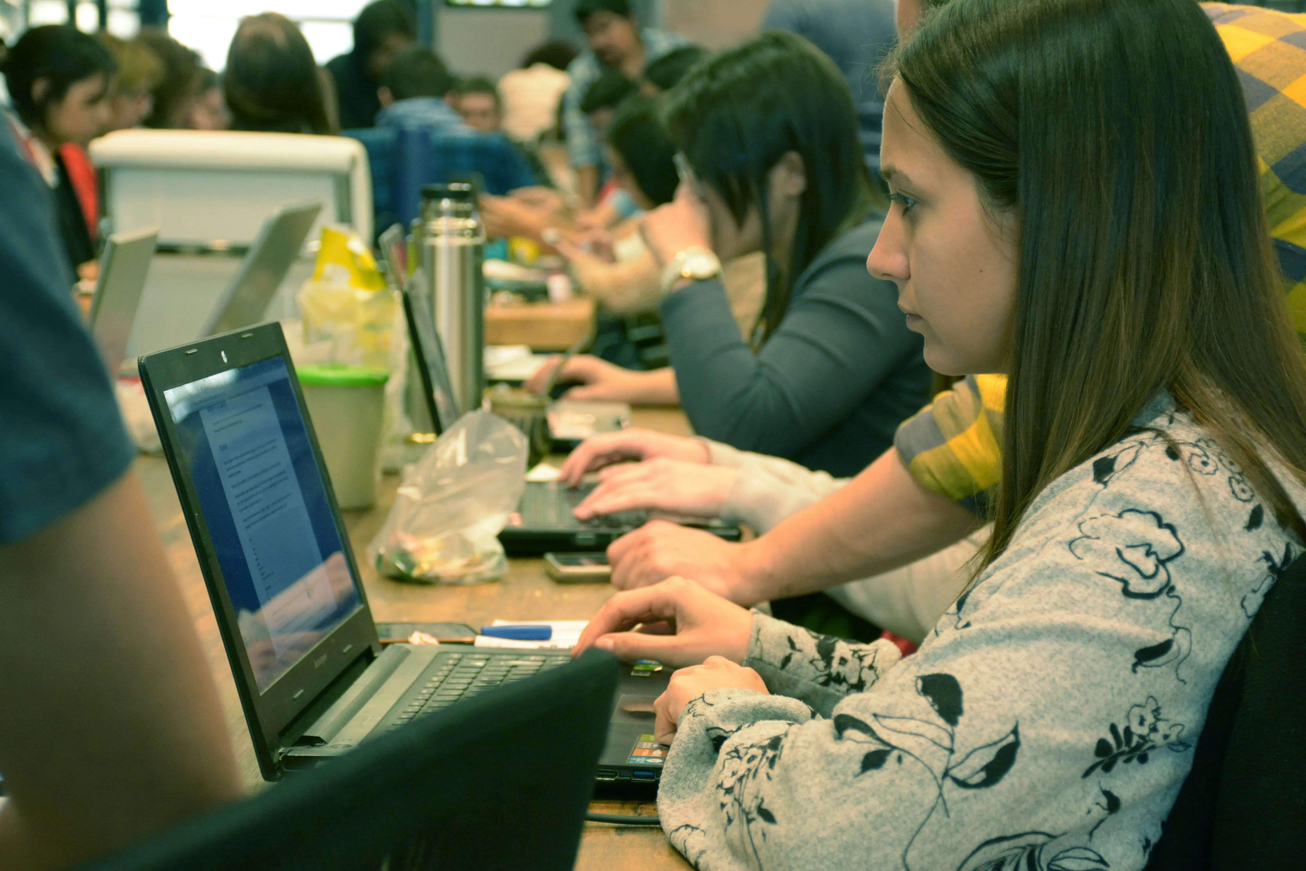 Young woman focused on typing on a laptop in a busy coworking space with people working in the background.