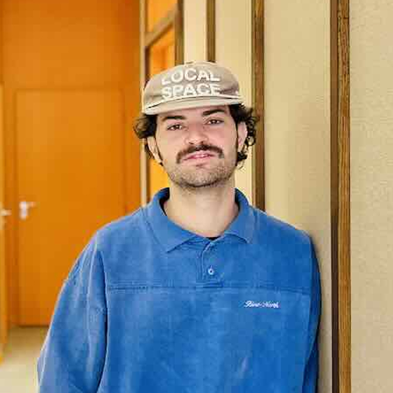 Portrait d'un homme aux cheveux courts bruns avec un léger sourire, en arrière-plan un salon lumineux avec des plantes et du mobilier moderne.