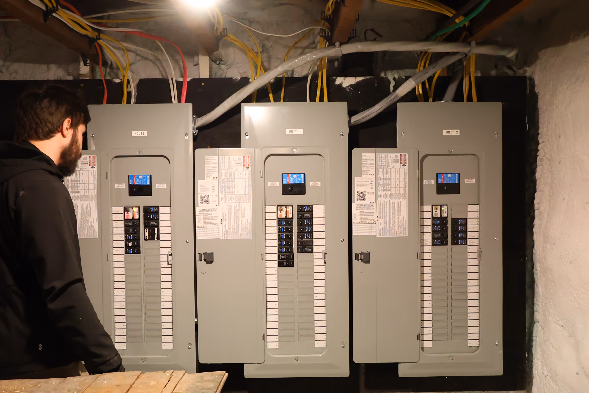Hall Electric inspecting three gray electrical breaker panels labeled House, Unit 1, and Unit 2 with various colored wires above them.