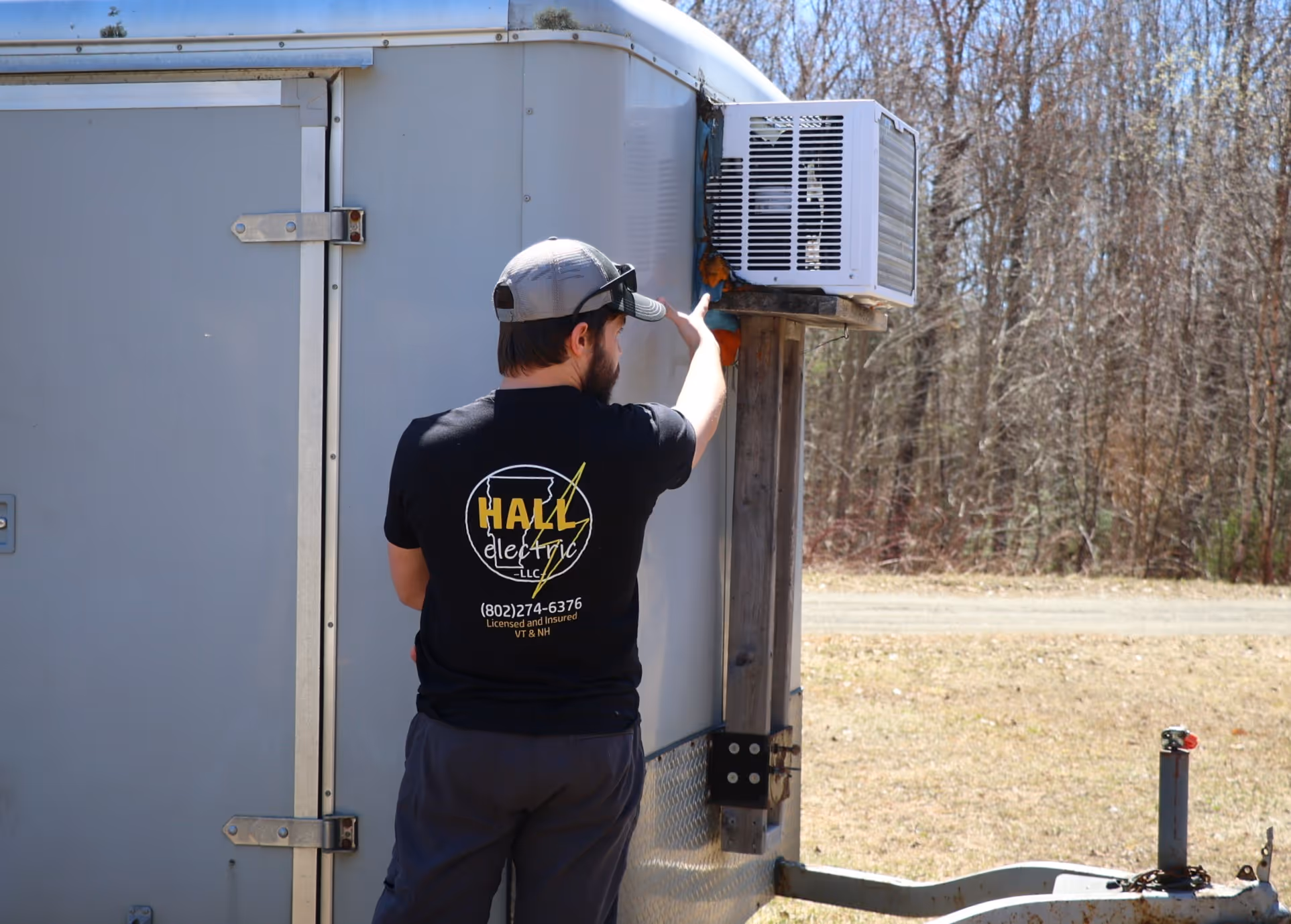 Man wearing Hall Electric t-shirt talking about an air conditioner attached to a trailer outdoors.