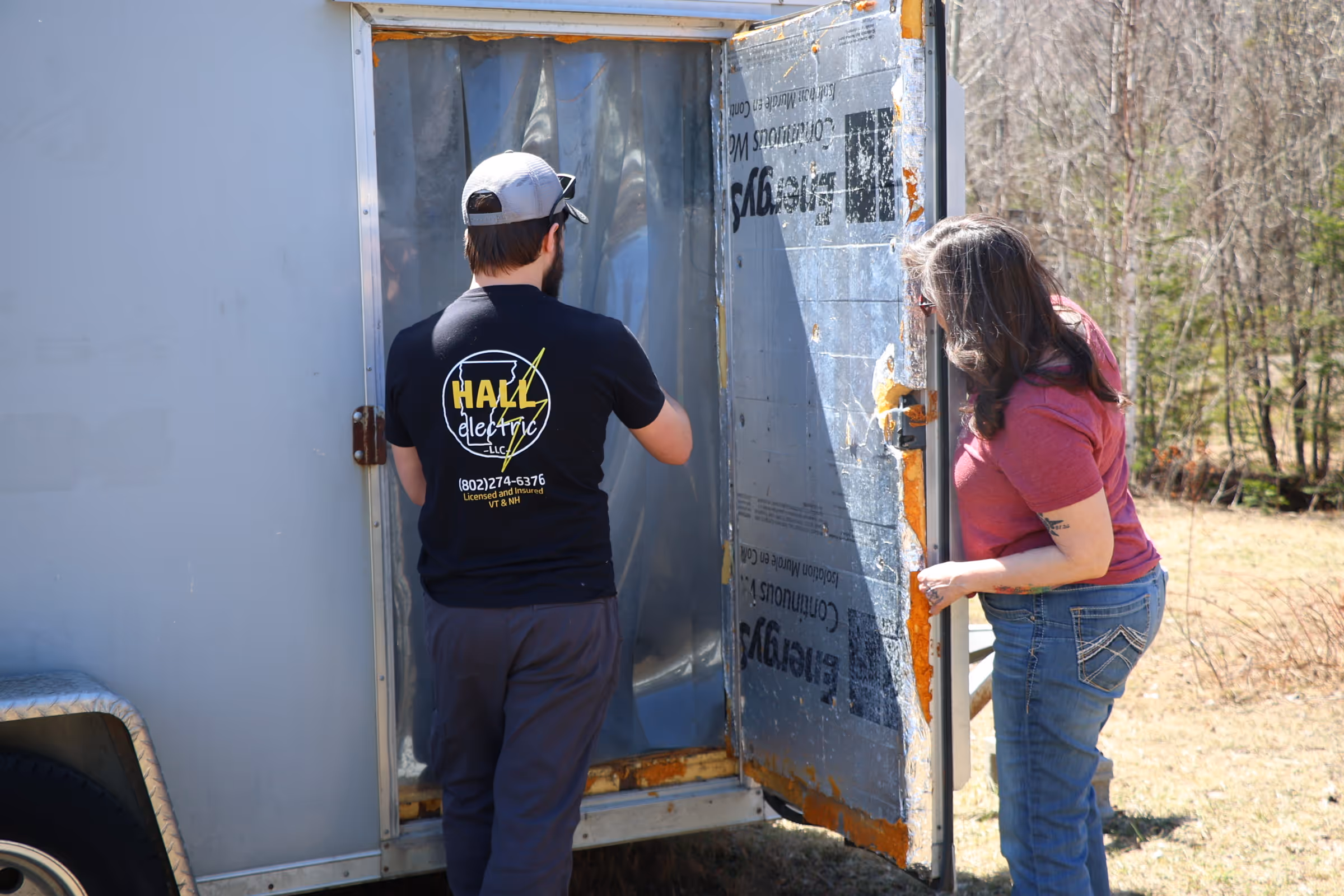 Man in a black Hall Electric shirt and woman in jeans inspecting the open door of a trailer outdoors.