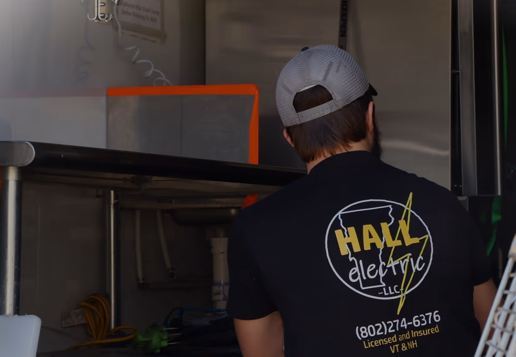 Man wearing a black Hall Electric LLC shirt with contact info and a gray mesh hat working on a food truck
