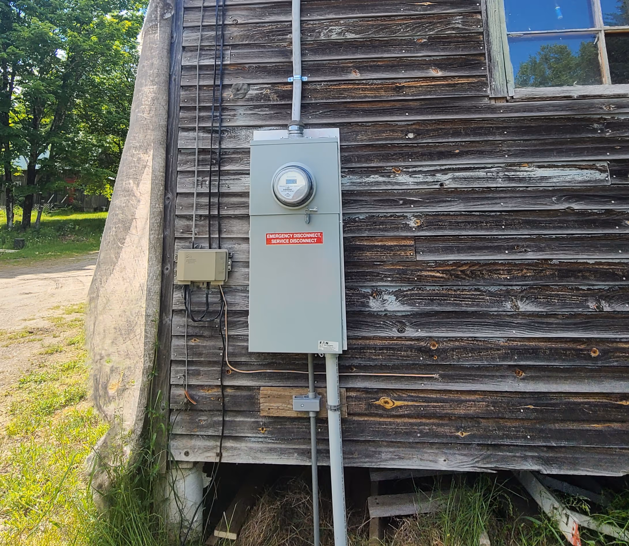 Gray electrical service disconnect box on aged wooden exterior wall with greenery nearby.