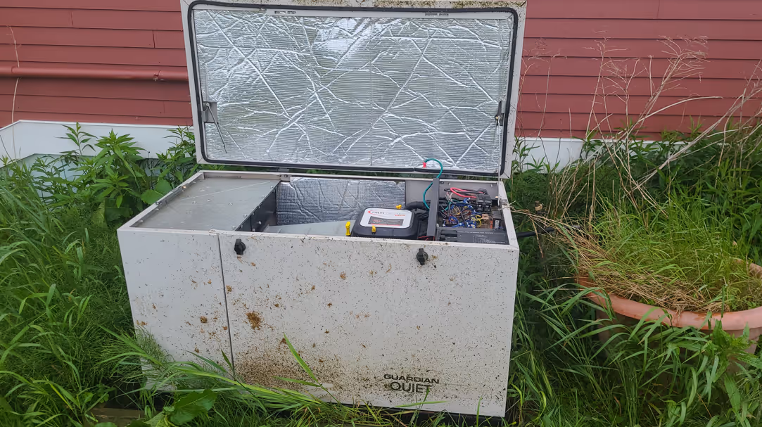 Old outdoor generator with open lid, showing internal components, surrounded by grass and plants.