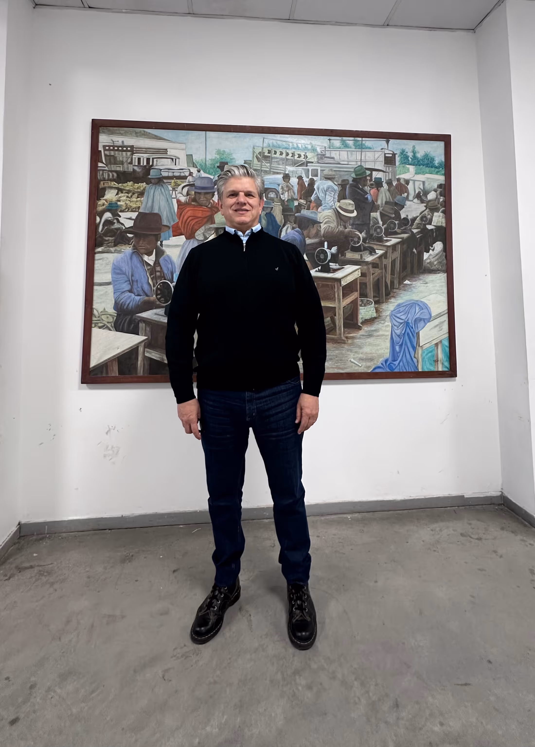 Hombre de pie frente a un mural que muestra varias personas trabajando en máquinas de coser en un taller al aire libre.