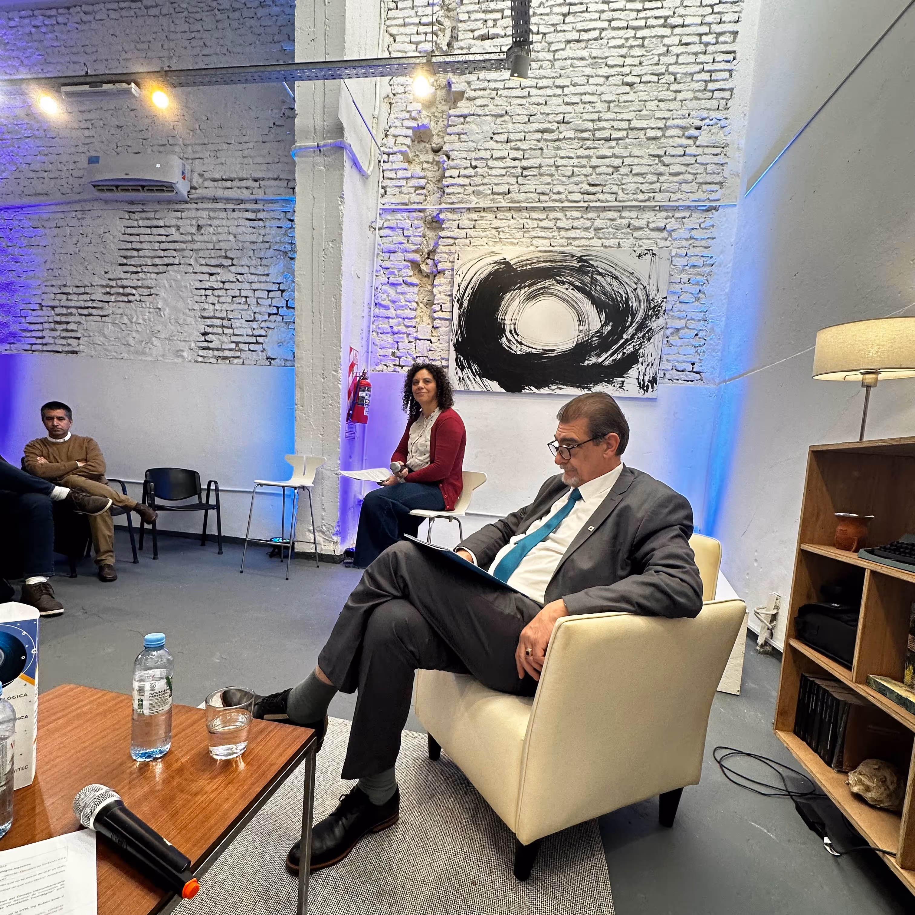 Dos personas sentadas en un espacio con paredes de ladrillo blanco; un hombre de traje y corbata sentado en un sillón beige y una mujer con suéter rojo y jeans sentada en una silla blanca, con una mesa con botellas, vaso y micrófono en primer plano.