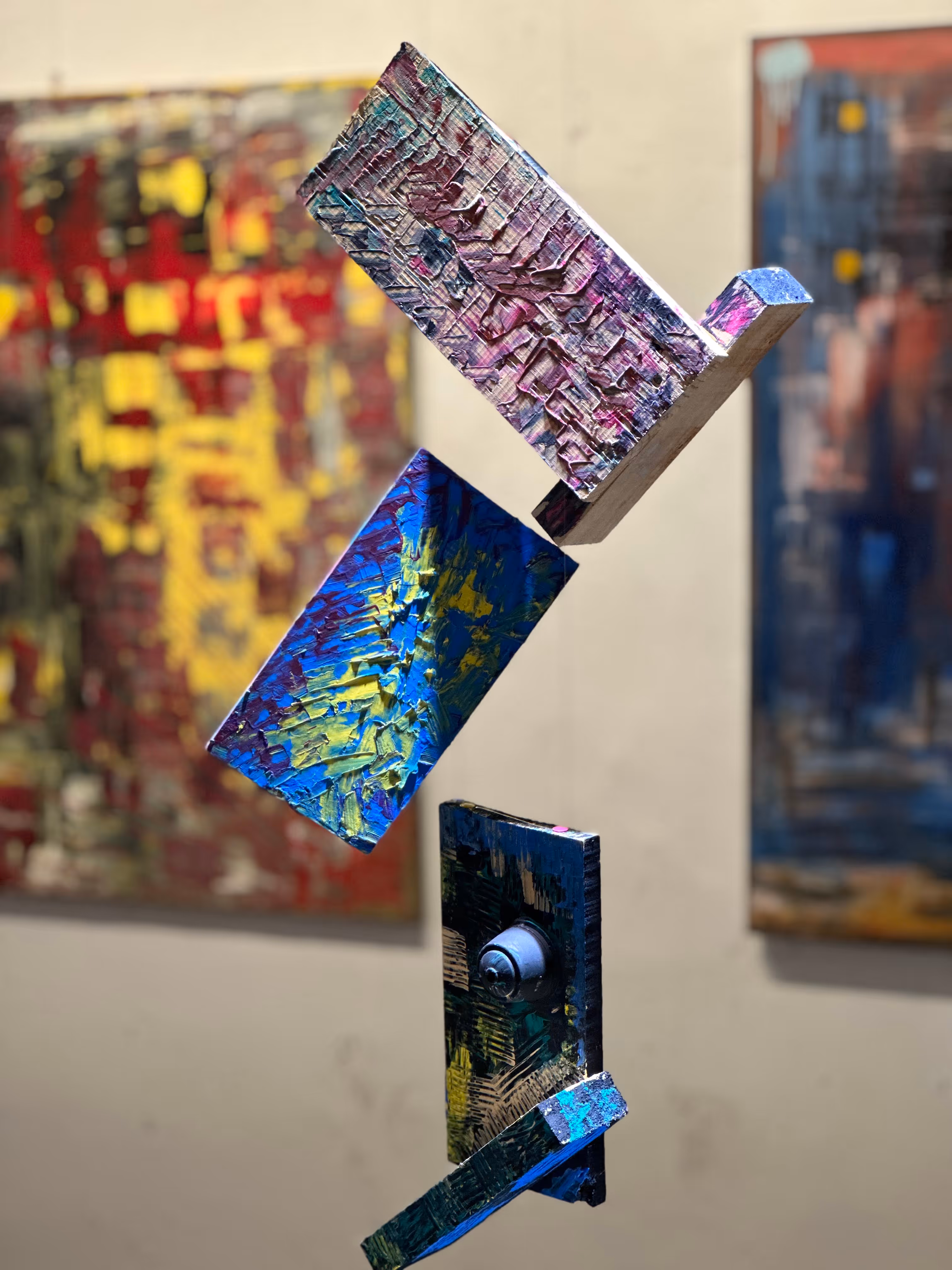 Escultura abstracta con placas de madera pintadas en colores vivos, suspendidas verticalmente con fondos de cuadros abstractos desenfocados.