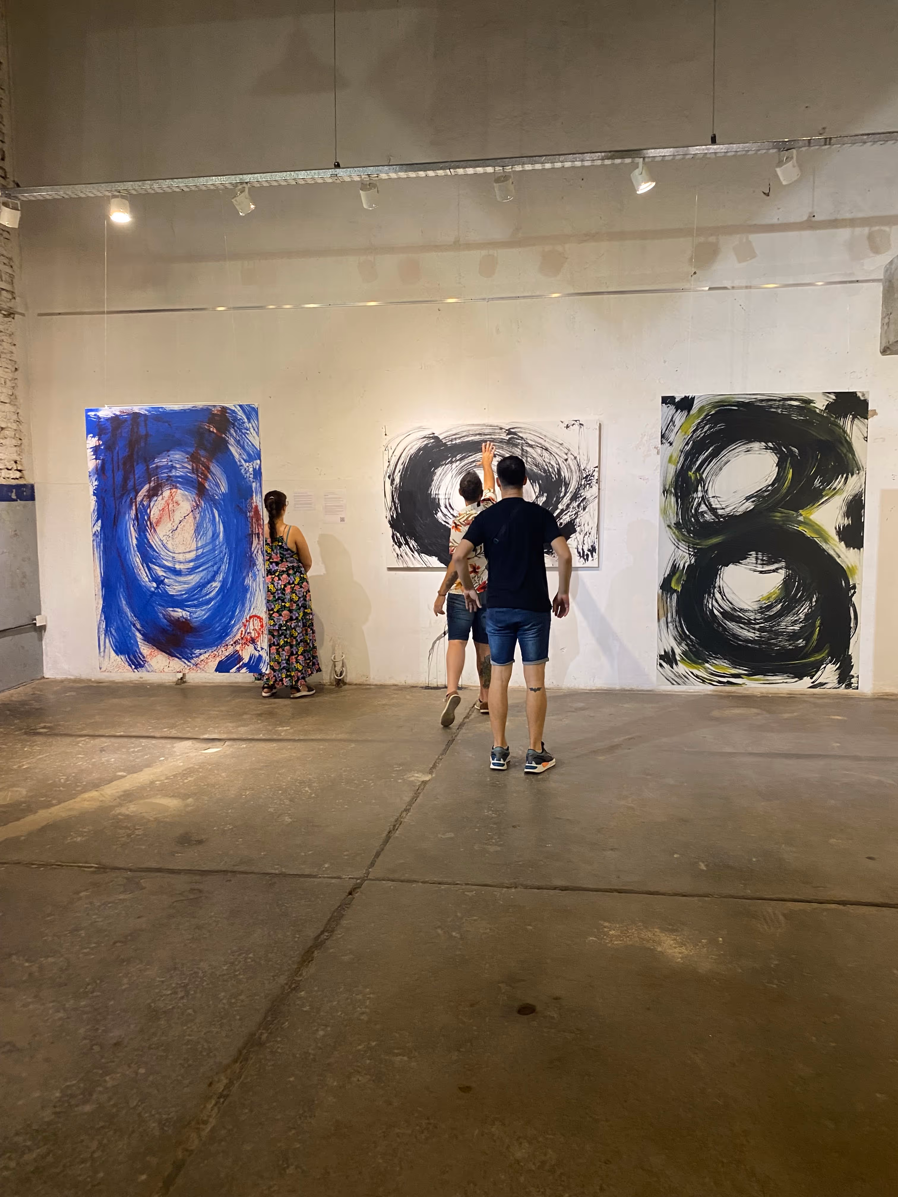 Tres personas observan pinturas abstractas con grandes círculos negros y azul en una galería de arte con paredes blancas y suelo de cemento.