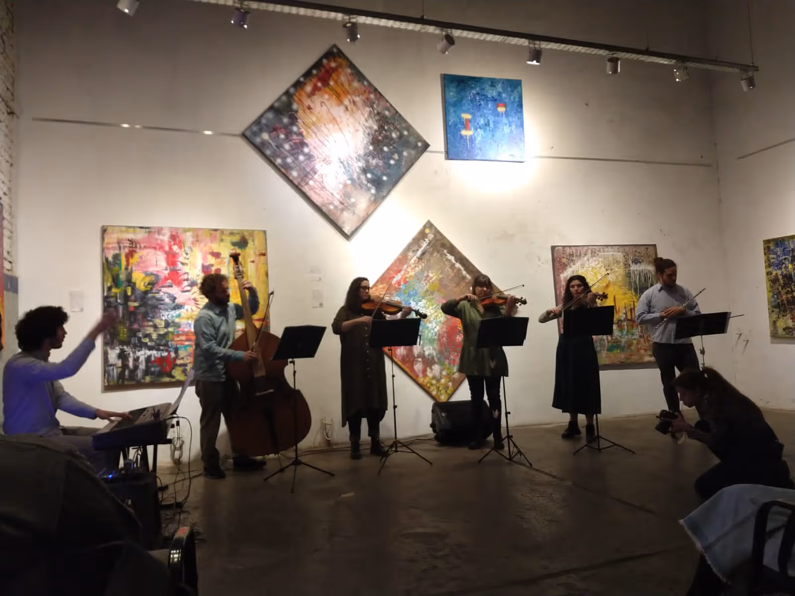 Conjunto de músicos tocando instrumentos de cuerda y teclado en una galería de arte con pinturas abstractas coloridas en las paredes.