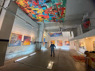 Galería de arte con cuadros coloridos colgados en muros blancos y un gran mural abstracto en el techo, con persona de pie en el centro.