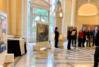 Personas de pie en un evento formal dentro de un edificio con paredes altas, columnas y obras de arte expuestas.
