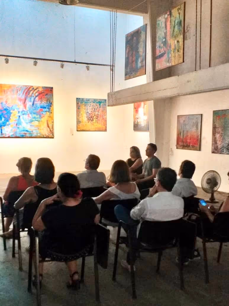 Personas sentadas en sillas dentro de una galería de arte mirando pinturas abstractas en las paredes.