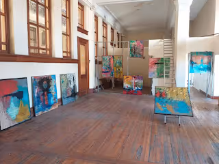 Dos hombres posan en una galería de arte con pinturas coloridas colgadas en las paredes y apoyadas en el suelo.