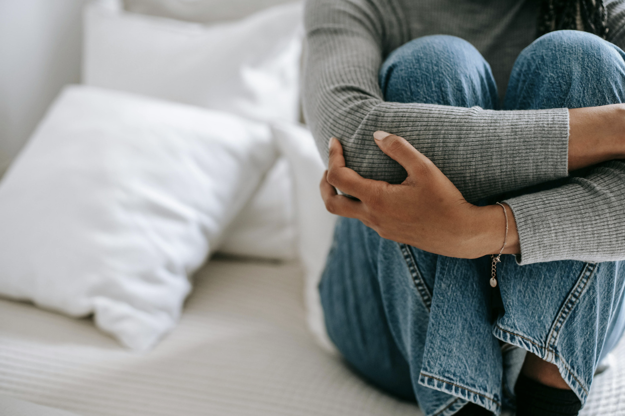 Person sitting on a bed with their knees drawn up, wearing jeans and a gray sweater, appearing anxious or distressed.