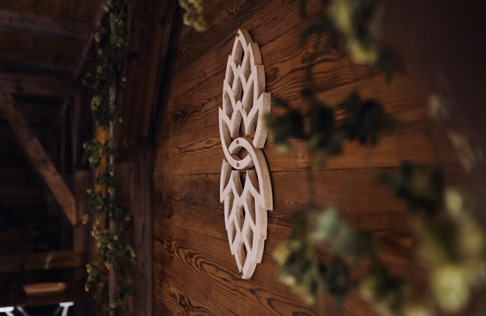 Holzwand mit einem geschnitzten, weißen Holzornament in Hopfenform, umgeben von Hopfenranken.