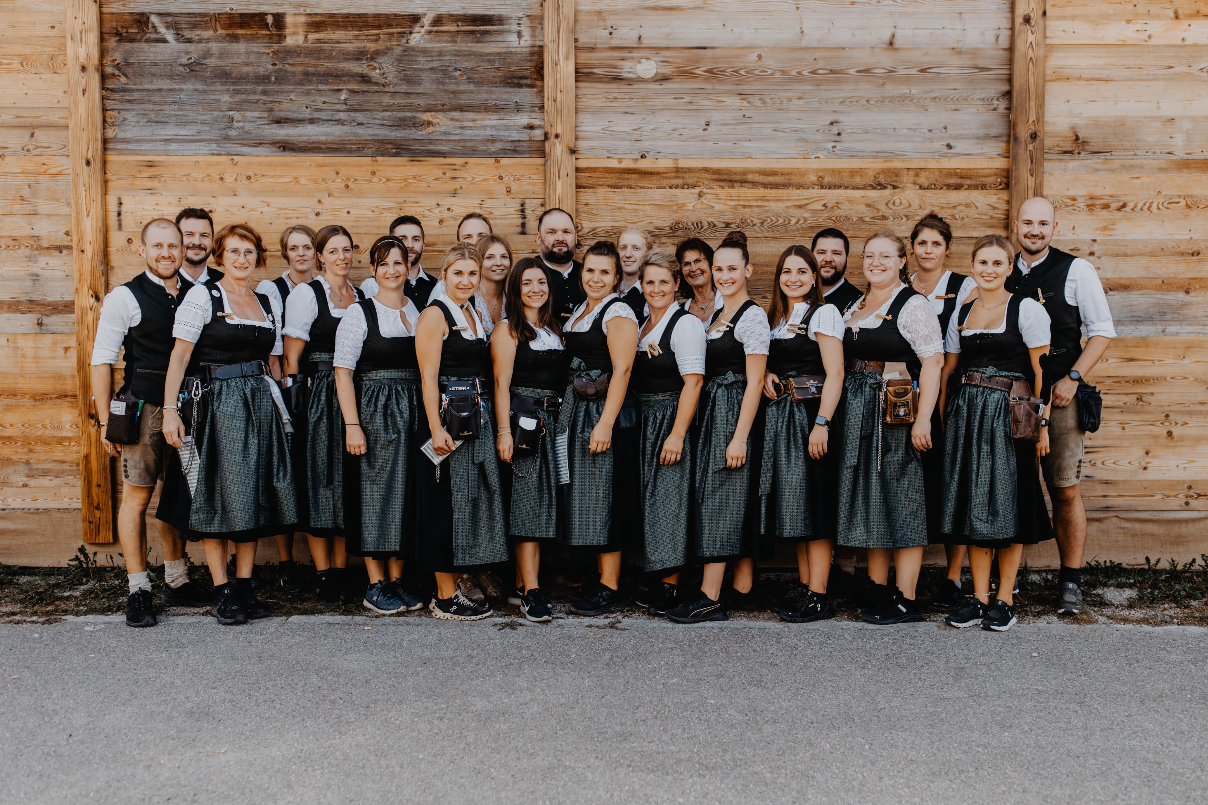 Gruppe von 25 Bedienungen des Erhartinger Festzelts in traditioneller bayerischer Tracht vor einer Holzfassade.
