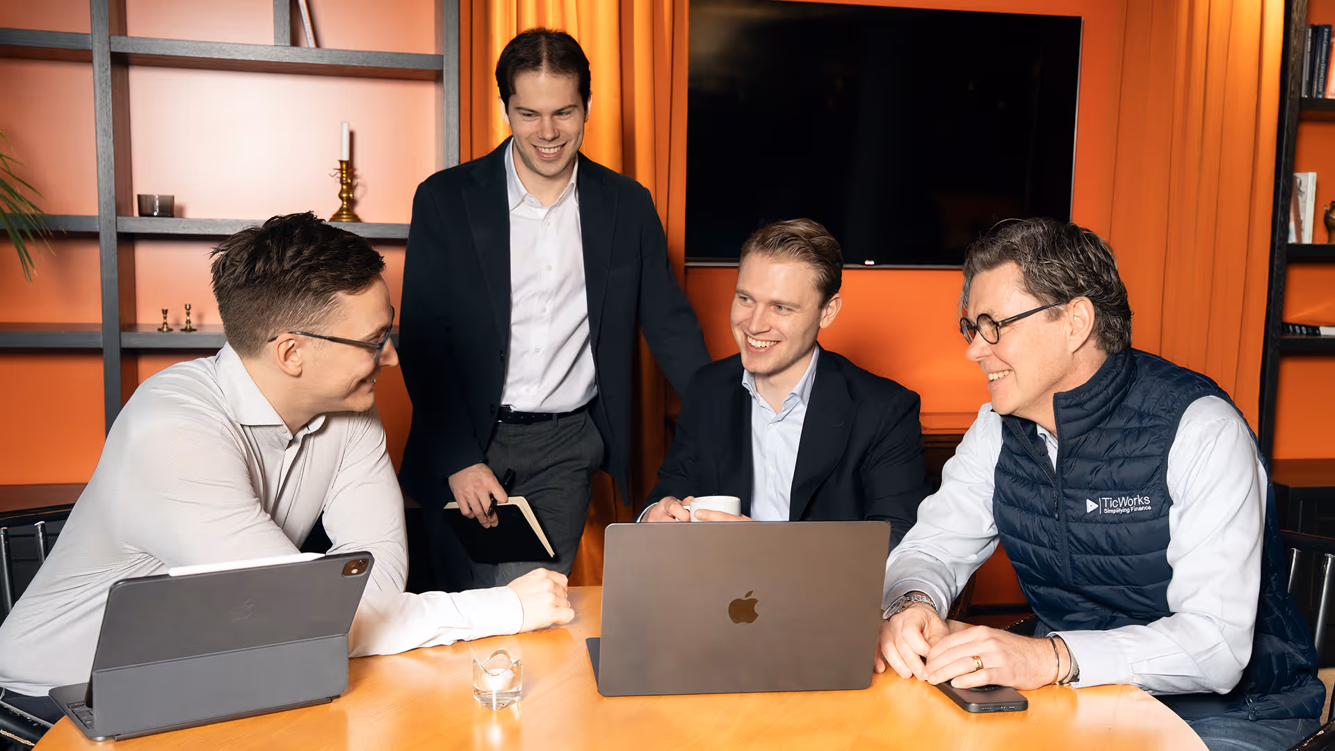 Fyra män i formella kläder diskuterar och ler runt ett bord med en laptop och surfplatta i ett kontorsrum med orange bakgrund.
