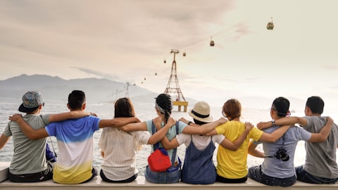 Groupe d'amis assis côte à côte avec les bras autour des épaules, face à la mer et à un téléphérique.