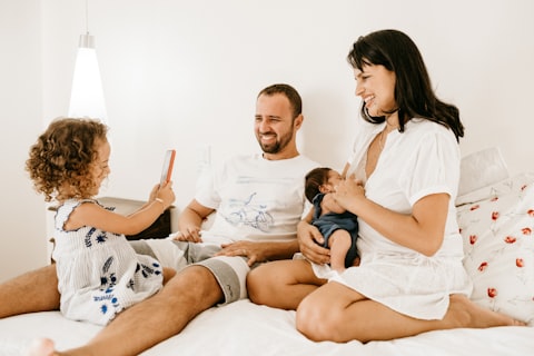 Famille souriante assise sur un lit, mère tenant bébé, père et fille regardant un téléphone.