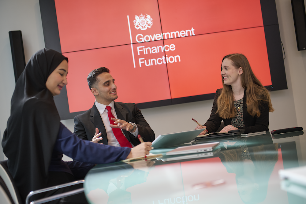 Three professionals having a discussion in a meeting room with a red screen behind them displaying 'Government Finance Function'.