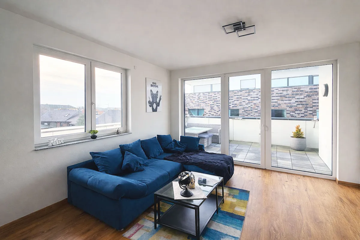Wohnzimmer mit blauem Ecksofa, Glastisch, Teppich, großen Fenstern und Zugang zum Balkon.