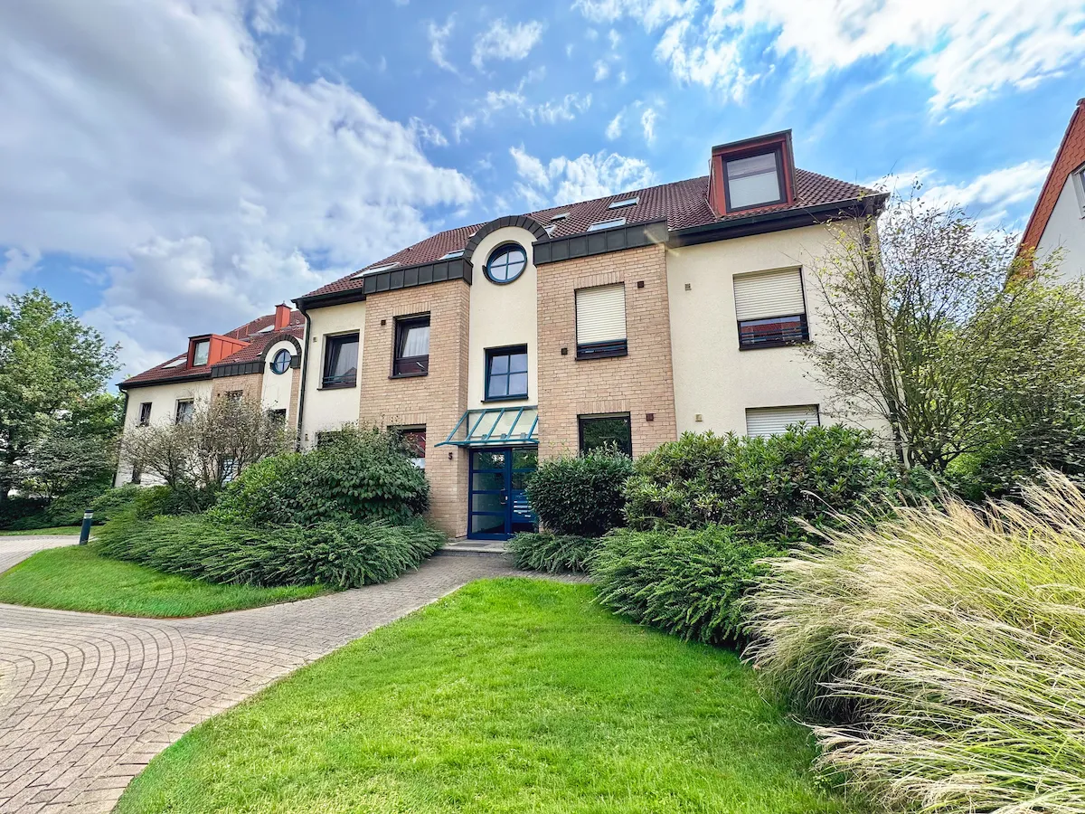 Mehrfamilienhaus mit gepflegtem Vorgarten, grünem Rasen und blauem Himmel mit Wolken.