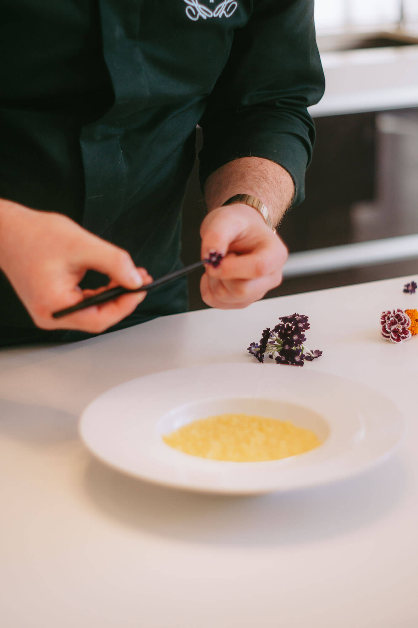 Chef decorates a plate of food with purple edible flowers using tweezers.