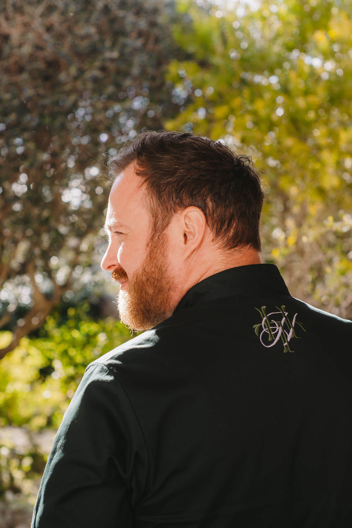 Bearded man in black shirt outdoors with embroidered logo on collar, looking to the left.
