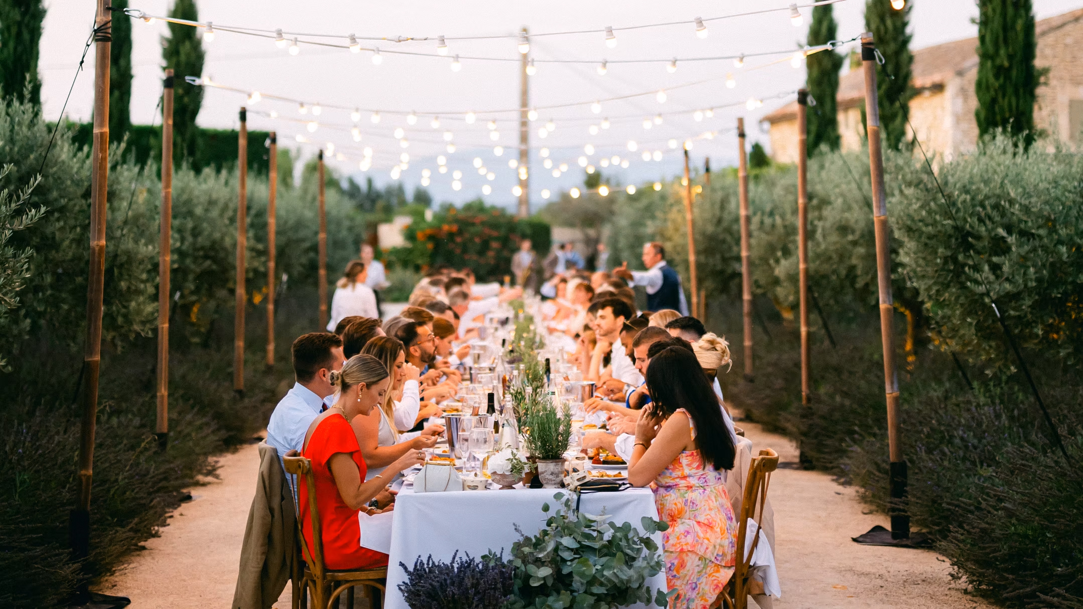 Long dining table with people seated outdoors under string lights in a garden setting at dusk.