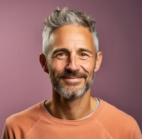 Smiling middle-aged man with gray hair and beard wearing a peach sweatshirt against a purple background.
