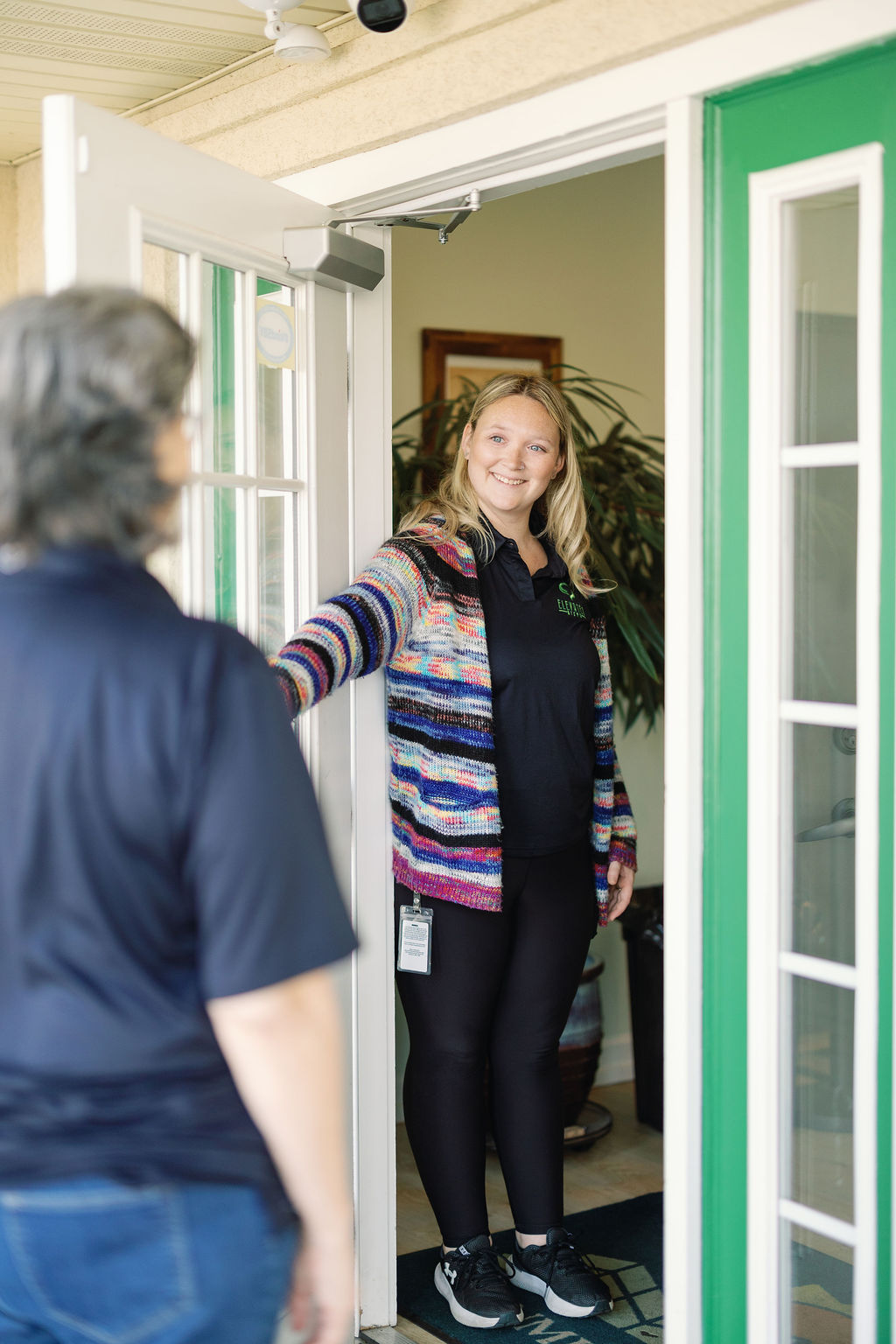 Woman with striped sweater holding door open, smiling to another person entering through a green door.