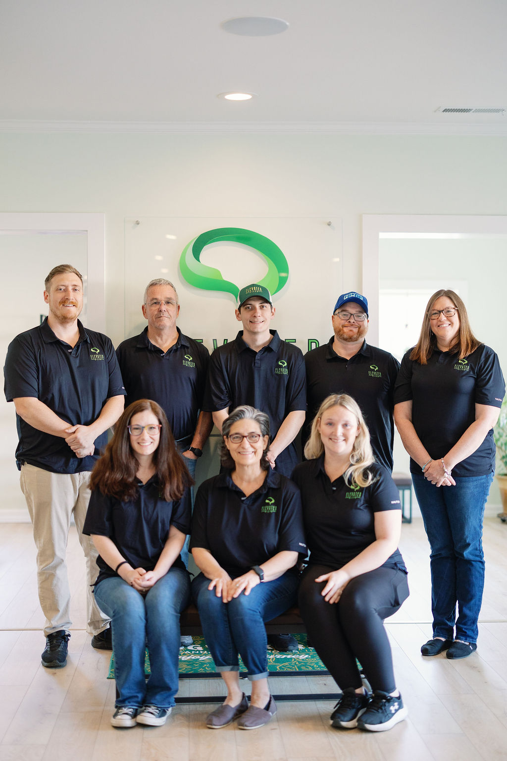 Eight people wearing matching black Elevated Landscaping shirts pose indoors in front of a company logo.