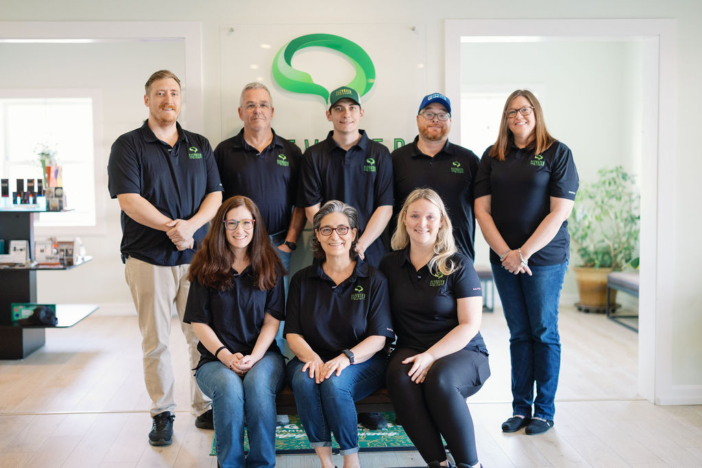 Group of eight people in black branded polo shirts posing indoors with a green logo behind them.